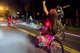 group of cyclists riding at night in an urban setting, wearing casual gear and lots of lights on the bikes