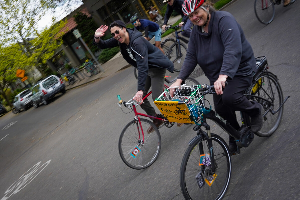 Two people smiling and riding bicycles on the street.