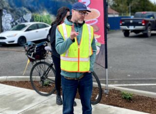Man in neon vest speaking while standing on sidewalk.