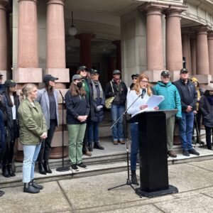 Group of people inside standing behind someone at a lectern in front of an old city building.