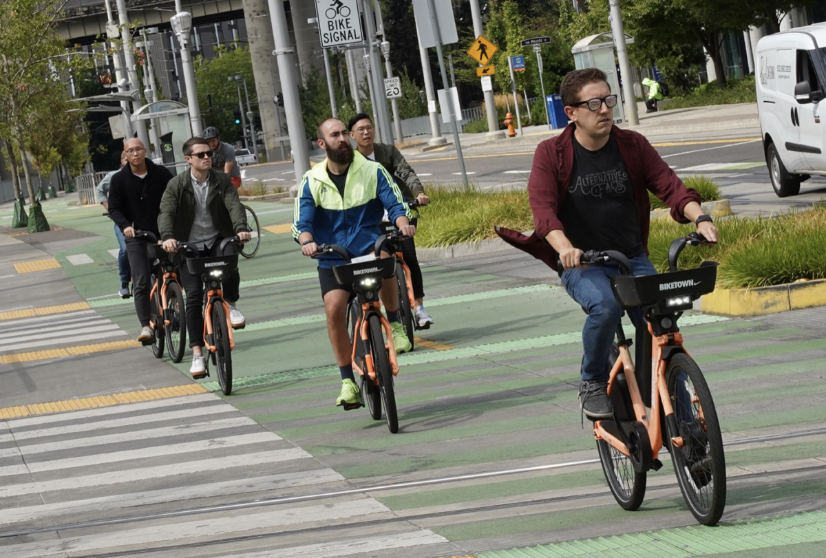 People riding orange shared bicycles on a green bike lane in an urban area.