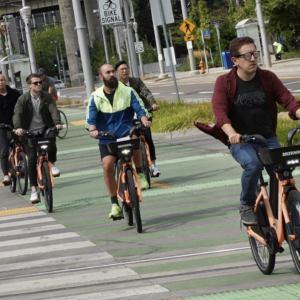 People riding orange shared bicycles on a green bike lane in an urban area.