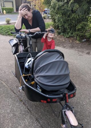 A mom with her head in her hands standing over a cargo bike with a baby seat in it.