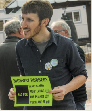 Freeway protester Aaron Brown carries a green sign with an anti-freeway message. He's wearing a shirt with two buttons on it and is speaking to someone out of frame.