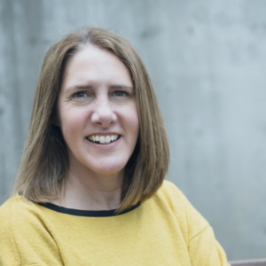 Portland of a smiling person with a yellow sweater and shoulder-length hair in front of a grey background.