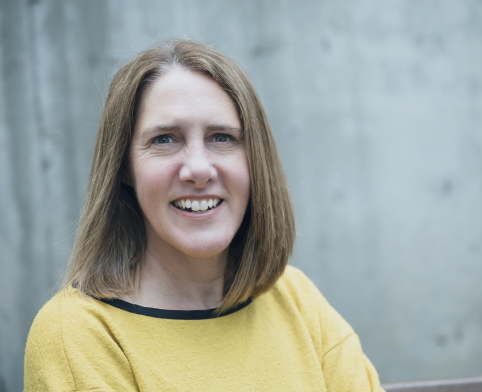 Portland of a smiling person with a yellow sweater and shoulder-length hair in front of a grey background.