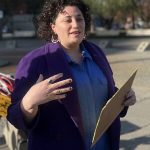 Person with curly hair and light brown skin making a hand gesture and holding a folder outside.