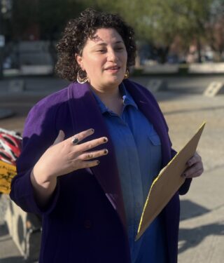 Person with curly hair and light brown skin making a hand gesture and holding a folder outside.