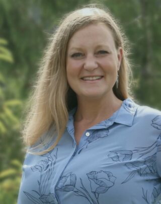 Person with blonde hair and blue shirt smiling in a portrait taken against a green outdoor background.