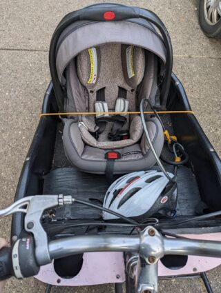Baby seat with a bungee cord across it inside the cargo bin of a bicycle.