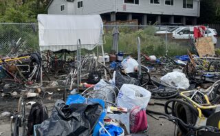 Piles of bikes and bike parts on a sidewalk next to a chainlink fence.