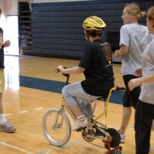 Person riding an adaptive bike in a gym with helpers running alongside them.