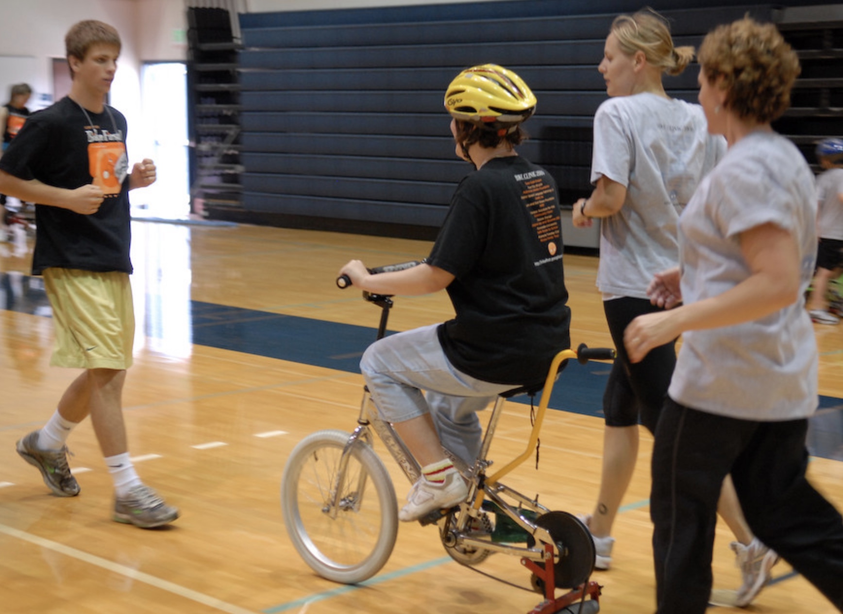 Person riding an adaptive bike in a gym with helpers running alongside them.