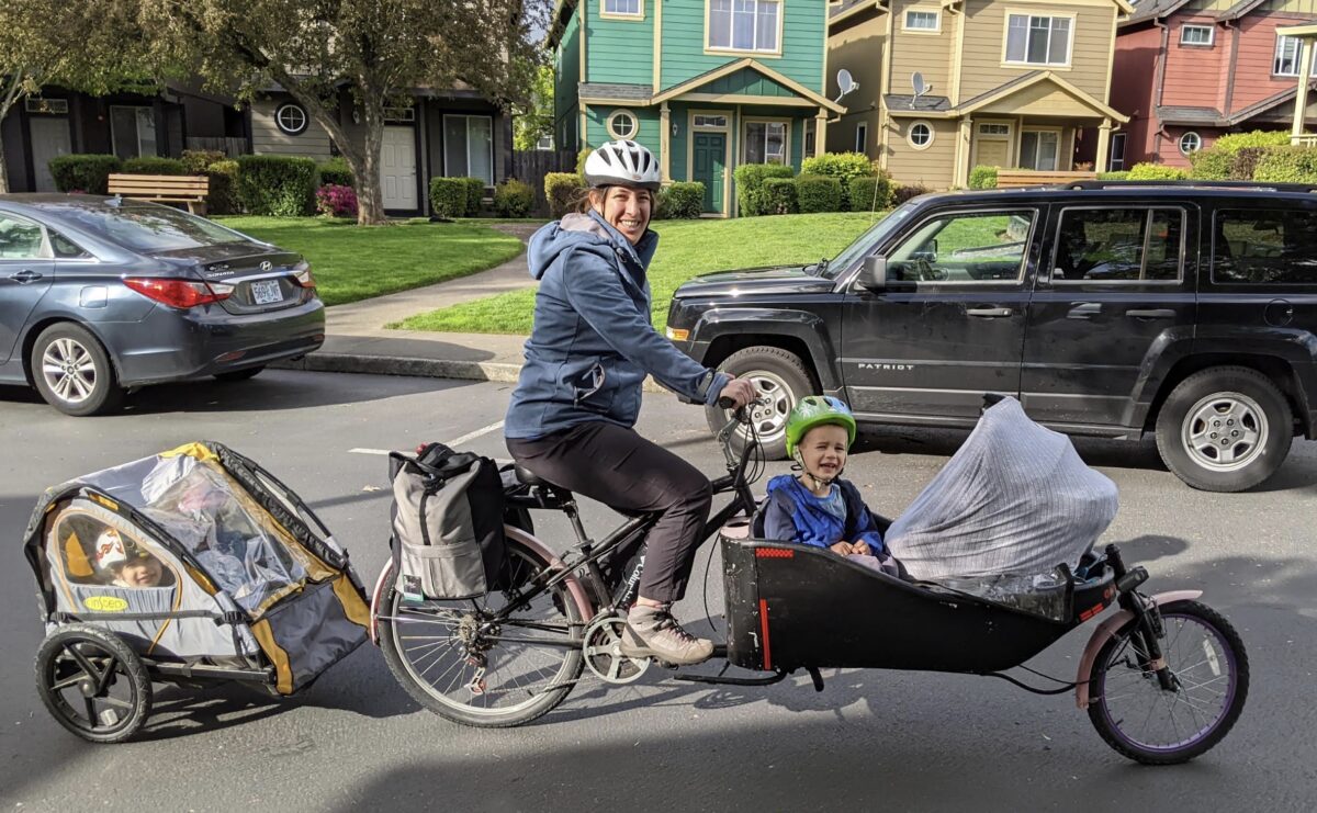 Person in the street on a bike pulling a child trailer and with children in a front cargo box smiling.