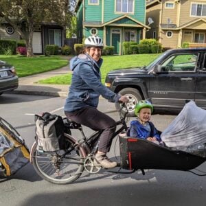 Person in the street on a bike pulling a child trailer and with children in a front cargo box smiling.