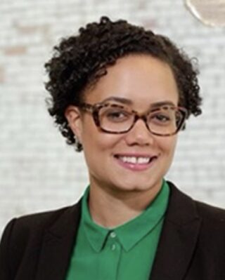 Portrait of a person with black curly hair and glasses standing in front of a white background