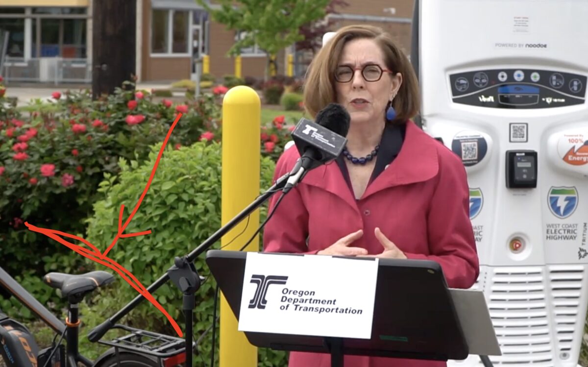 Oregon Gov Kate Brown speaking outside at a press event in a red shirt in front of an EV charging station with a bike nearby.