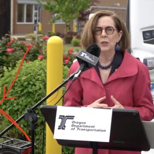 Oregon Gov Kate Brown speaking outside at a press event in a red shirt in front of an EV charging station with a bike nearby.