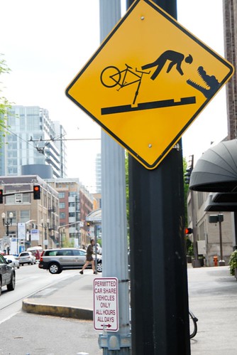 Yellow street sign with an illustration of a person riding a bike crashing into an alligator, artwork added later by a street artist.